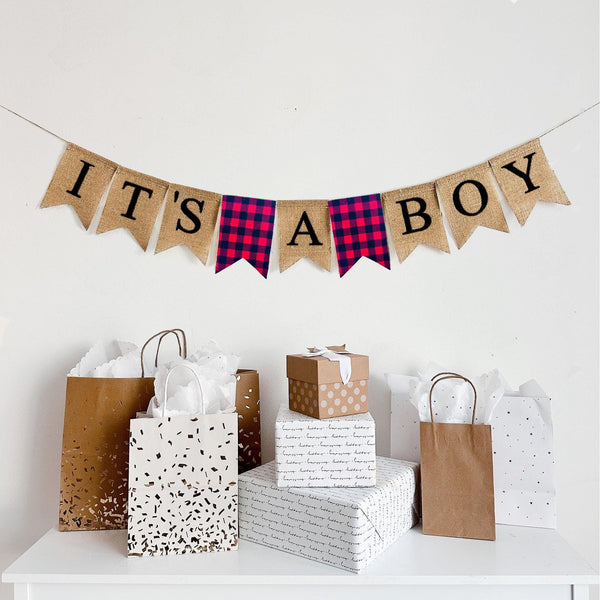 A burlap banner made from 4.5 by 6.5 inch fishtail shaped flags. Each flag has a pocket across the top for stringing. This banner says IT&#39;S A BOY in black capital letters and has fishtail shaped flag of red and black check fabric between each of the words. It is strung on a 110 inch jute string.