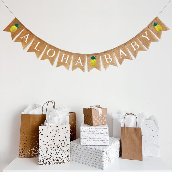 An Aloha Baby burlap banner featuring three hand stenciled pineapple image flags at the beginning, middle and end of the banner. Each fishtail burlap flag measures 4.5 x 6.5 inches with white lettering. The pineapple image flags are at the beginning, middle and end of the banner making this banner a tropical and festive addition to your event.