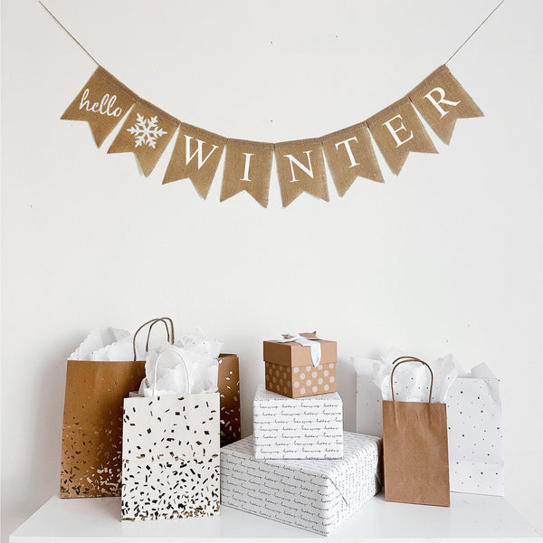 A Hello Winter burlap banner. Printed on burlap fishtail flags, this banner hangs on one line. The first flag says &quot;hello&quot; stenciled in white paint in script lettering. The next flag is a white glittered snowflake followed by flags spelling out the word WINTER in white letters. Each burlap flag measured 4.5 x 6.5 inches.