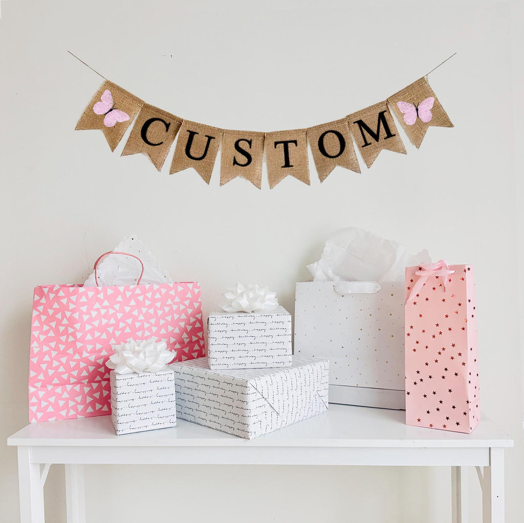 A burlap banner made from 4.5 by 6.5 inch fishtail shaped flags. Each flag has a pocket across the top for stringing. Add a custom name or word of your choice in black capital letters. There is an image of a pink butterfly on each end of the word. It is strung on a 110 inch jute string.