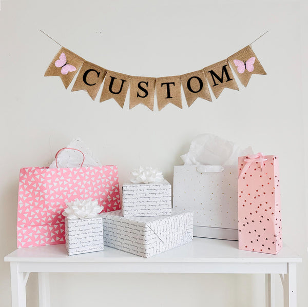 A burlap banner made from 4.5 by 6.5 inch fishtail shaped flags. Each flag has a pocket across the top for stringing. Add a custom name or word of your choice in black capital letters. There is an image of a pink butterfly on each end of the word. It is strung on a 110 inch jute string.