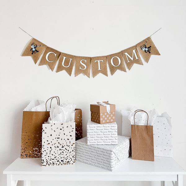 A burlap banner made from 4.5 by 6.5 inch fishtail shaped flags. Each flag has a pocket across the top for stringing. Add a custom name or word of your choice in white capital letters. There is an image of a monkey swinging on a vine on each end of the word. It is strung on a 110 inch jute string.