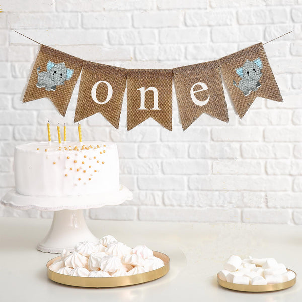 one first birthday banner with grey elephants with blue ears on each end. the banner spells out o n e in lowercase white letters on burlap flags.