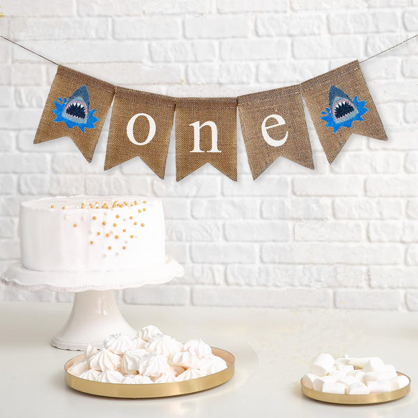 A burlap banner that says one with a shark icon on each side.
