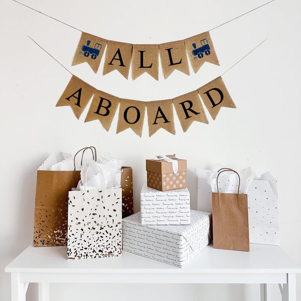 A two tiered burlap banner that says all aboard in black lettering. The first tier says all with a navy blue train flag on either side of the word. The second tier says aboard.