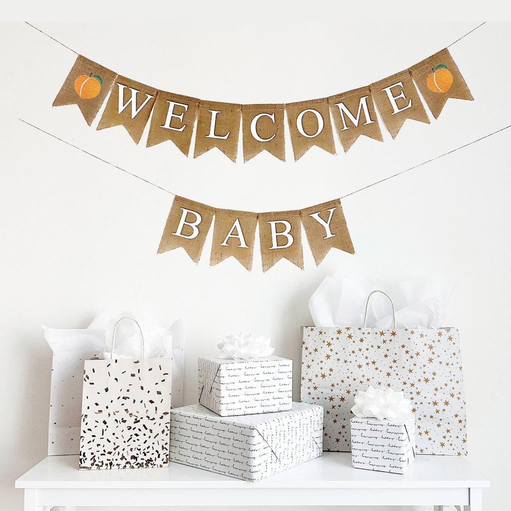 A burlap baby shower banner that reads Welcome Baby in white font. Featuring two stenciled peaches on either end of the word welcome with the word baby on the second line, this banner is the perfect backdrop to a summer baby shower.
