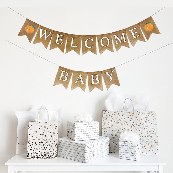 A burlap baby shower banner that reads Welcome Baby in white font. Featuring two stenciled peaches on either end of the word welcome with the word baby on the second line, this banner is the perfect backdrop to a summer baby shower.