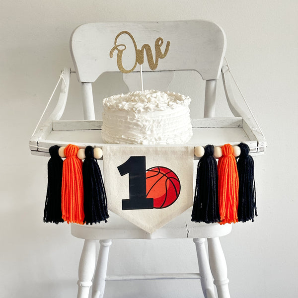 May include: White high chair with a white cake on top. The cake is decorated with white frosting and has a gold glitter topper that says &#39;One&#39;. A banner with a basketball and the number &#39;1&#39; is hanging from the high chair. The banner is decorated with orange and black tassels.