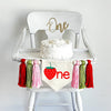 May include: White high chair with a white cake on top. The cake is decorated with white frosting. A banner with a red strawberry and the word &#39;one&#39; is hanging from the chair. The banner is decorated with pink, green, and red tassels.