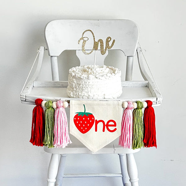 May include: White high chair with a white cake on top. The cake is decorated with white frosting. A banner with a red strawberry and the word &#39;one&#39; is hanging from the chair. The banner is decorated with pink, green, and red tassels.