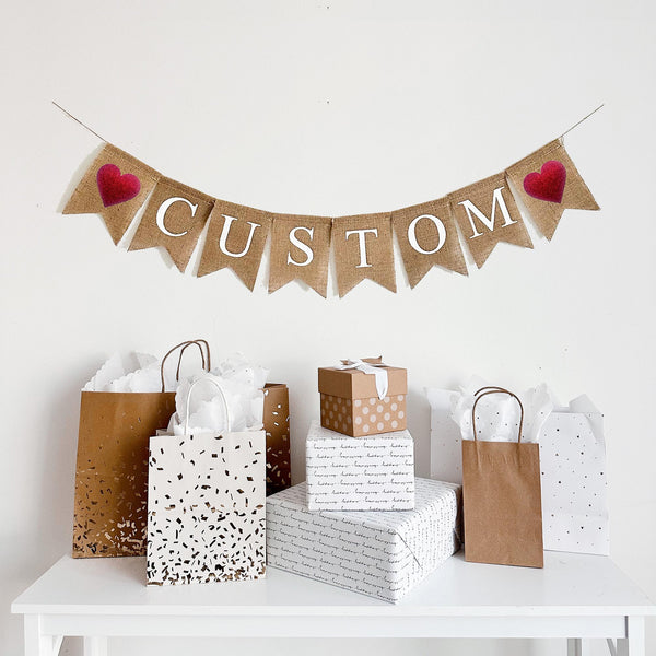 A burlap banner. The flags say custom- you can add any text you&#39;d like. There is a red glitter heart on each end of the word. The letters are white and all capitals.