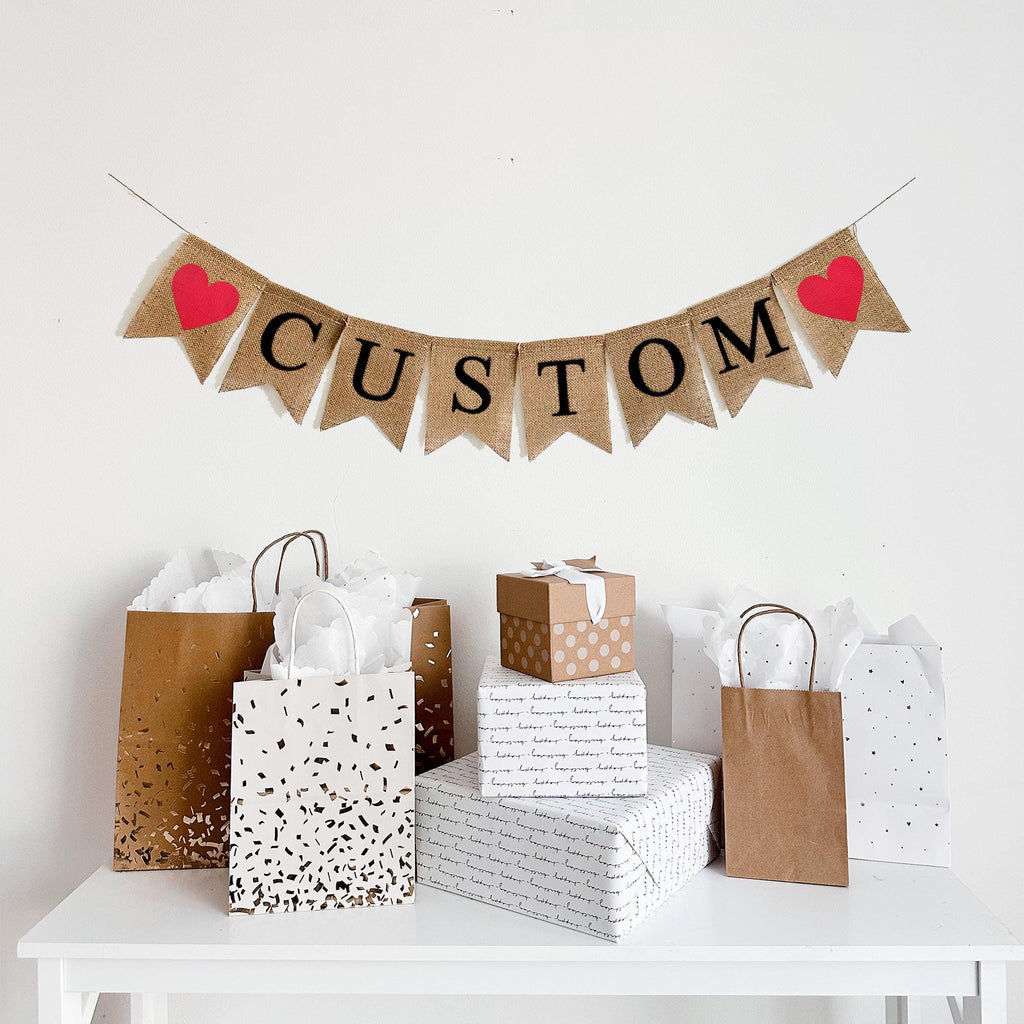 A burlap banner. The flags say custom- you can add any text you'd like. There is a red glitter heart on each end of the word. The letters are black and all capitals.