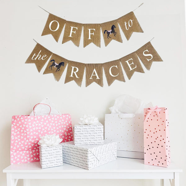 A burlap banner made from 4.5 by 6.5 inch fishtail shaped flags. Each flag has a pocket across the top for stringing. The top string of this banner says OFF in white capital letters and TO in white script one on flag with a hand stenciled image of a horse with a pink saddle in between the words. The bottom string says THE in white script on one flag and RACES  in white capital letters with a hand stenciled horse with pink saddle in between the words. It is strung on two 110 inch jute strings.