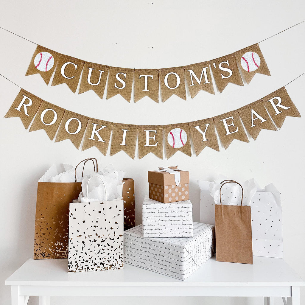 A burlap banner made from 4.5 by 6.5 inch fishtail shaped flags. The top line can be customized with a name in white letters and has a baseball image on each end. The bottom line says ROOKIE YEAR in white and has a baseball image in the middle.