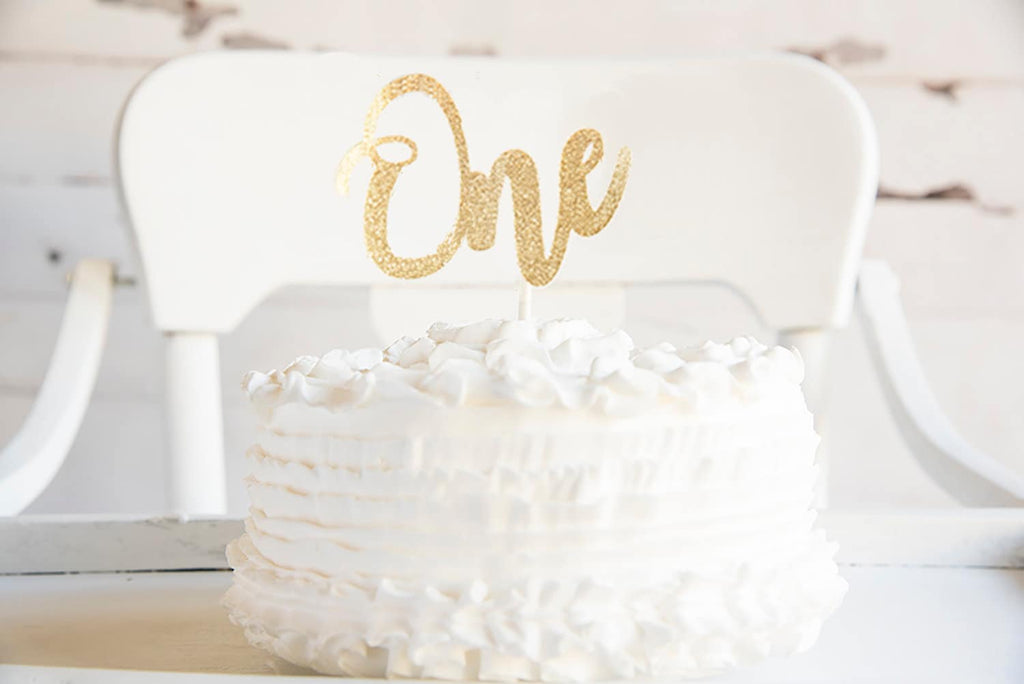 A white cake with a gold glitter One cake topper on it.