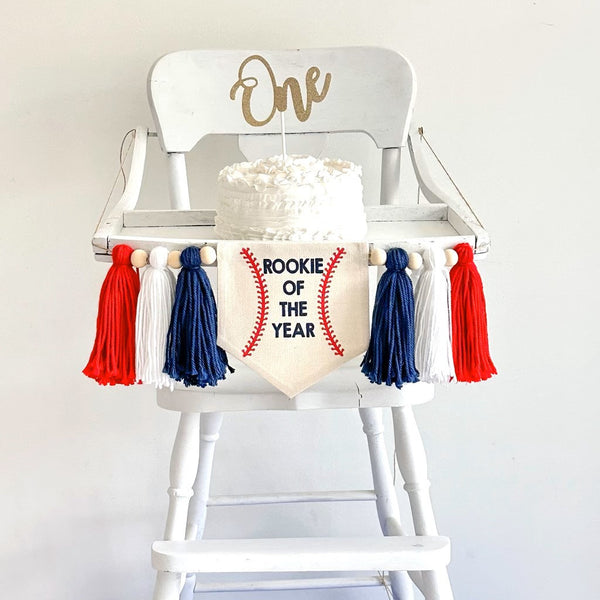 Rookie of the year banner with tassels and wooden beads attached to a highchair that has a cake with a one cake topper.