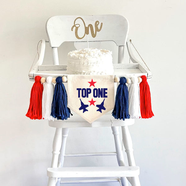 The wording top one is on a banner with 2 fighter jets in navy blue and with 2 red stars. The banner is attached to a highchair with yarn tassels. A cake with a one cake topper is on the highchair table. The color scheme is red, white and blue.